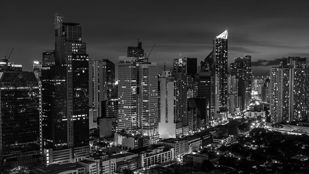 Black and white photo of a city skyline at night with illuminated buildings.